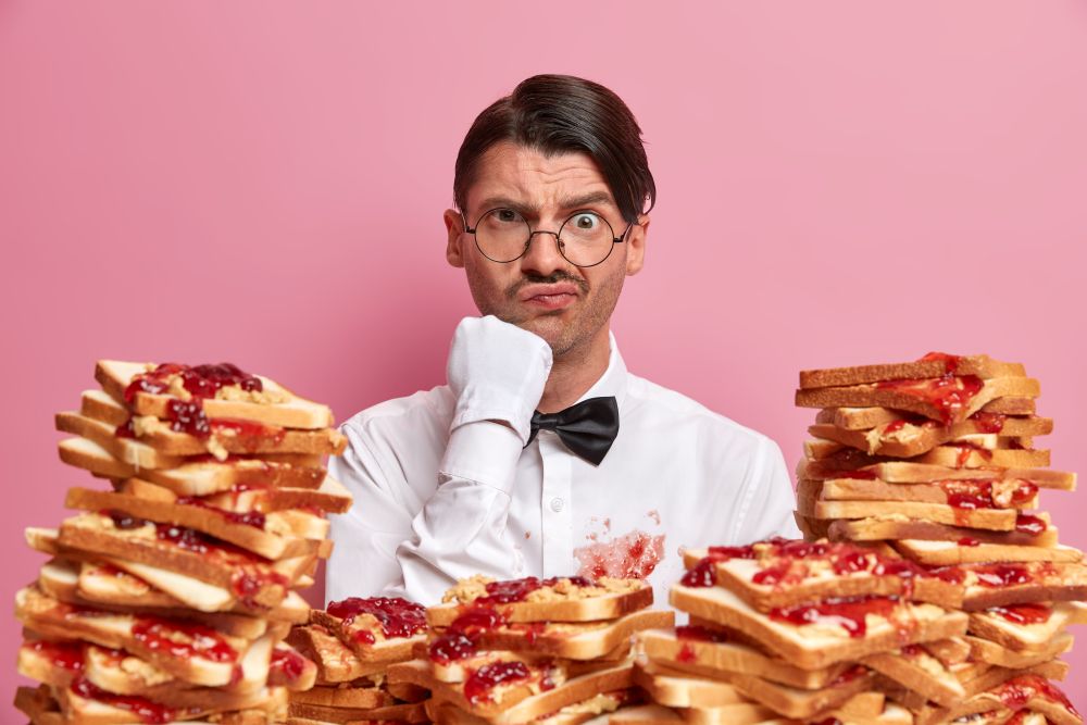 Serious man aristocrat frowns face, dressed in snow white elegant clothes, stands near stack of delicious snacks, isolated on pink background. Cafe staff indoor. Job position. Best waiter service